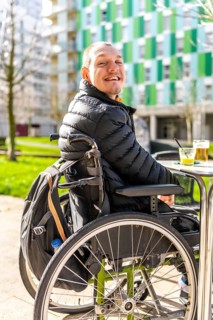 Happy disabled man sitting in a wheelchair, enjoying a refreshing drink at an outdoor table in an