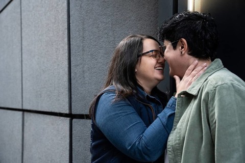 A curvy lesbian couple enjoys an intimate moment, smiling and embracing in an urban setting. They
