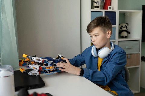 A young boy focuses intently on a robotic hand project at his desk. Wearing a blue shirt and