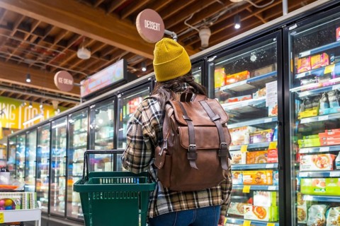 Woman in beanie and plaid shirt with backpack and shopping basket browsing frozen food in a modern