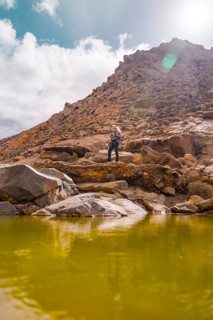 A woman explores rocky terrain beside a vibrant green mountain pond, with rocky cliffs and a blue