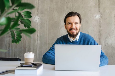 A professional man in a blue sweater smiles while working on a laptop at a modern desk. The office