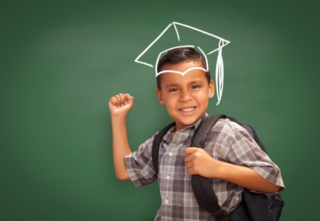 Young Hispanic Student Boy Wearing Backpack Front Of Blackboard with Graduation Cap Drawn In Chalk