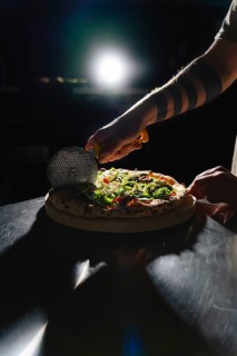 A hand with a tattoo slices a gourmet pizza topped with arugula, cheese, and cherry tomatoes using