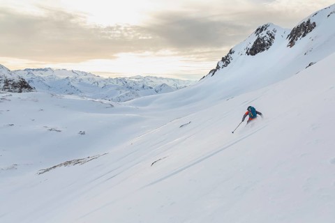 A lone ski tourer skis down a pristine, snow-covered mountain slope under a bright, cloud-streaked