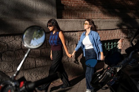 A multiethnic lesbian couple joyfully holding hands as they walk together on a sunlit street. They
