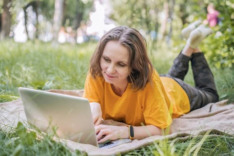 A woman lying on a blanket in a sunny park, using a laptop. She appears focused, combining work