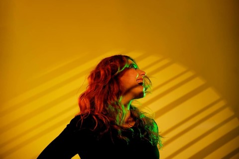 A young cheerful woman with flowing hair is surrounded by vibrant green and yellow studio lights.
