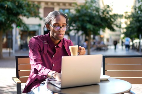 A man in a casual setting enjoys a cup of coffee at an outdoor cafe. He sits with his laptop,