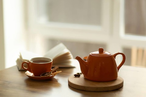 A cozy setup featuring an orange teapot on a wooden tray, a matching cup with tea, and an open book