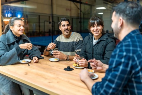 Group of young friends relaxing, drinking coffee and tea, conversing, and smiling around a table at