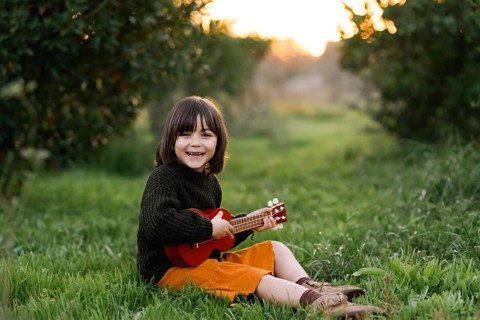 A cheerful child sits in a grassy meadow, strumming a small red guitar. The vibrant scene captures