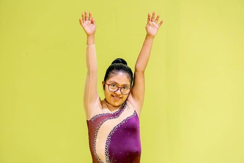 A young disabled gymnast stands confidently with arms raised against a bright green backdrop. She