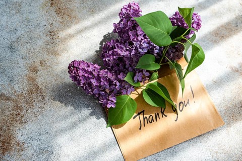 A charming bouquet of lilac blooms rests atop a handwritten thank you note on a rustic background.