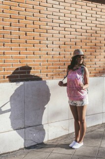 A stylish woman wearing a hat and sunglasses stands by a brick wall, casting a shadow. She is