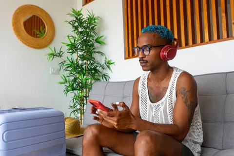 LGBTQIA+ man relaxes on a couch, using a smartphone and headphones to plan a holiday. A suitcase