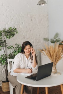 Nutritionist sitting at a white table, engaged in a call with a client. She's smiling, taking notes