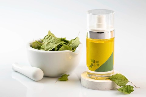 A clear bottle of birch oil stands beside a white mortar and pestle filled with green leaves, set