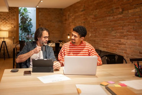 Two coworkers engage in discussion at a modern coworking space. One holds a tablet and coffee, the