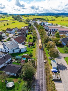 Aerial view of a village with railway line, surrounded by green fields and various houses, tamping