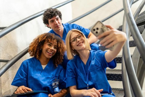 Three smiling medical students in blue scrubs are taking a cheerful selfie together on a metal