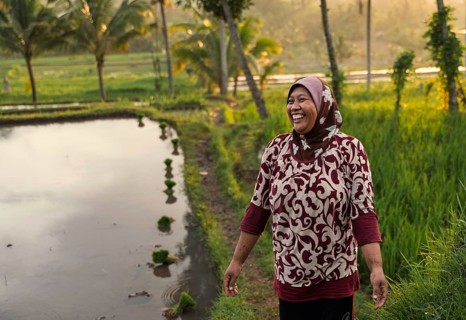 A smiling woman stands by a rice paddy in Indonesia, embraced by the warm glow of a sunset. Palm