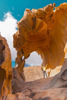 A woman wearing a yellow jacket stands triumphantly beneath a large, naturally formed rock archway,