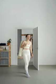 A woman in white active wear enters a yoga studio, carrying a mat bag over her shoulder. She exudes