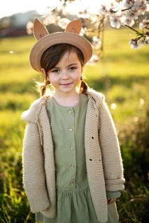 A young girl, dressed in a green dress and cozy sweater, stands in a field with almond blossoms in