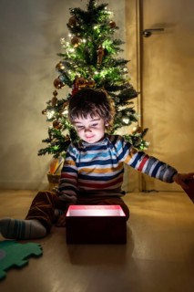 A young boy is sitting on the floor in front of a Christmas tree, opening a red box