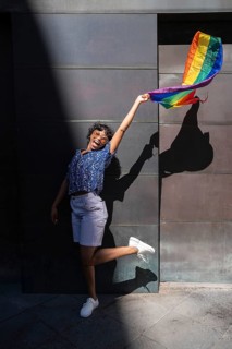 A joyful LGBTQ+ person wearing sunglasses holds a vibrant rainbow flag, casting a shadow against a