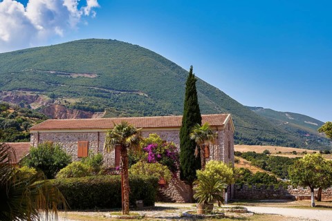 Rustic stone house in front of wooded hill under blue sky, surrounded by green vegetation, Achaia