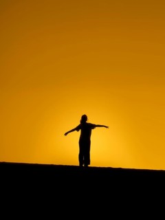 A woman stands on a hill in Fuerteventura at sunset, arms outstretched against a stunning orange