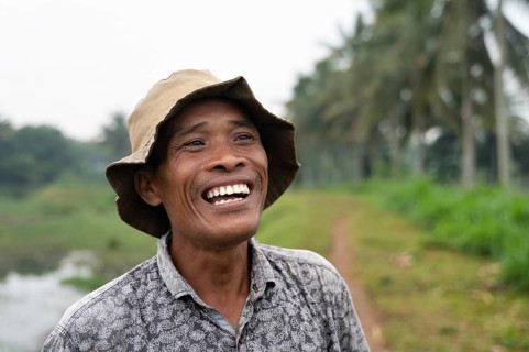 A joyous Indonesian farmer stands in a lush rice field, wearing a hat and patterned shirt. The