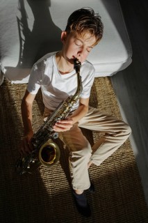 A young musician sits on a carpet, eyes closed, playing a saxophone. Sunlight creates a shadow,