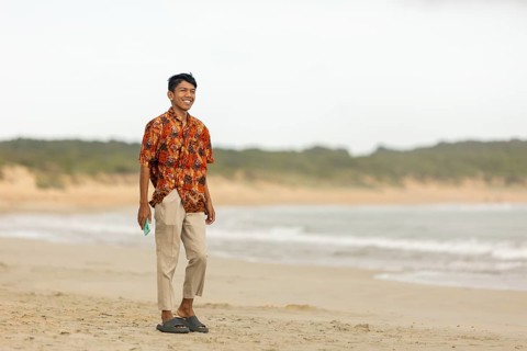 Smiling Indonesian man wearing vibrant batik shirt, walking along an Australian beach. Sandy shore,