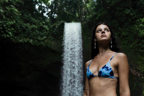 A woman in a blue bikini stands in front of a majestic waterfall surrounded by lush greenery,