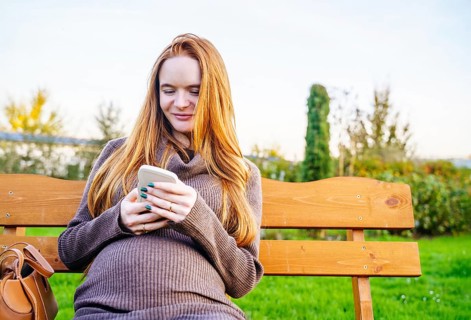 A beautiful pregnant woman with vibrant red hair is seated on a wooden bench in a serene outdoor