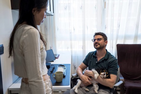 A diverse couple shares a warm moment at home, one seated with a small dog in an office chair,