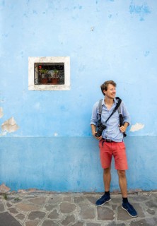 Young man in front of blue house wall with camera, small window with red flower, Burano Island,