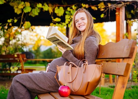 A serene pregnant woman with beautiful red hair sits on a wooden bench in an autumn park, engrossed