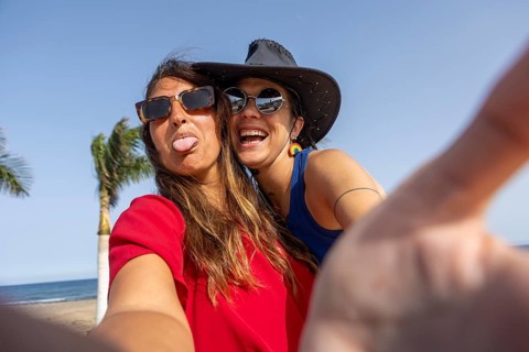 A cheerful lesbian couple takes a selfie on a sunny beach, capturing the carefree vibe of summer.
