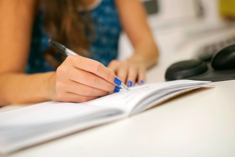 A close-up shot of a hand with blue nails writing with a pen on paper. The setting is an office