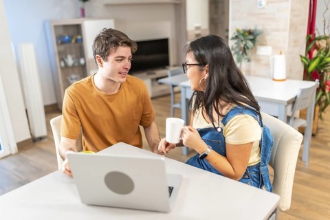 Young multiethnic couple enjoying coffee and working on a laptop at their kitchen table, sharing a