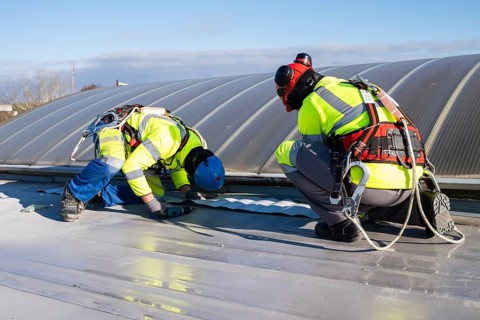 Two roofers wearing helmets and harnesses are carrying out sealing work on a flat roof near a