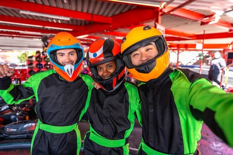 Diverse friends wearing helmets and racing suits, smiling and taking a selfie together at a go kart