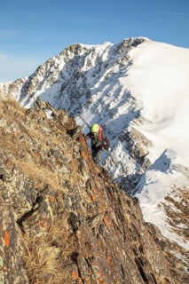 A ski tourer navigates a rocky mountain ridge, surrounded by snow-capped peaks under a clear blue
