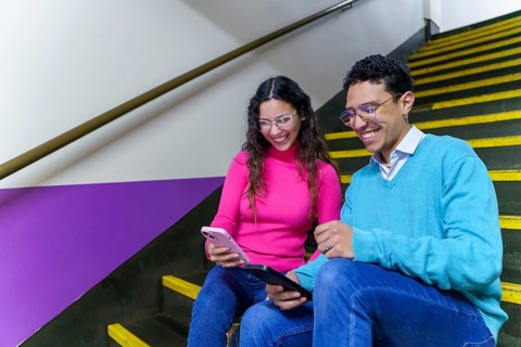 A cheerful smiling young woman and man, both Gen Z, smile while using a smartphone and tablet on a