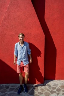 Young man leaning against a red house wall, colourful houses on the island of Burano, Venice,