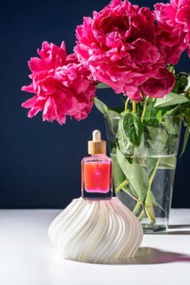 A vibrant pink serum in a dropper bottle is elegantly placed on a white marble stand. Lush peonies